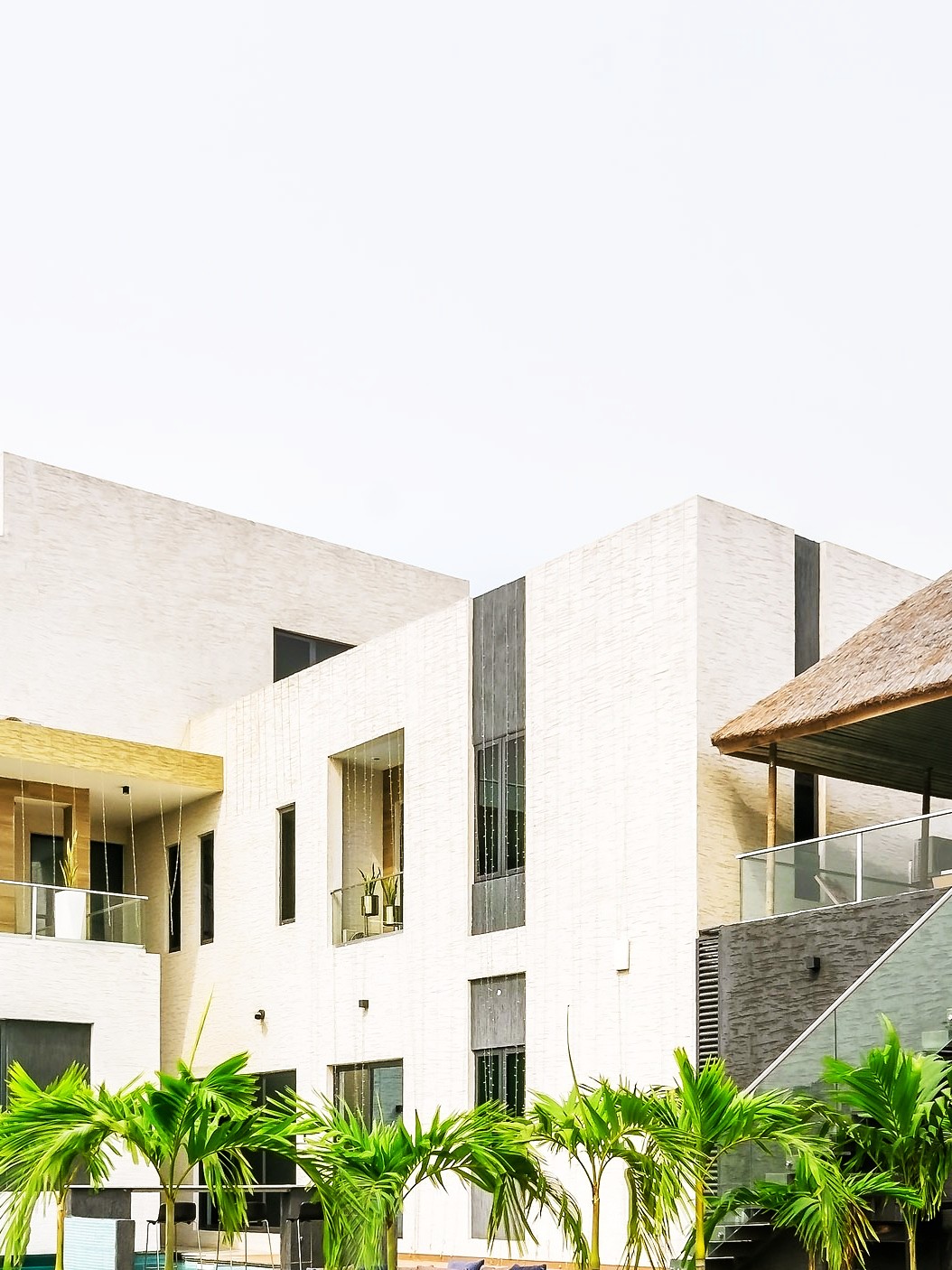 Elevated view of a contemporary white residence with a swimming pool, timber pool deck, thatched pavilion, landscaped lawn and perimeter fencing.